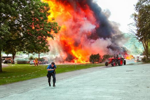 Firefighter in blue shirt and dark pants stands in foreground as massive orange and black flames erupt from a structure in the distance.