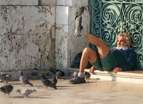 Man sitting on ground surrounded by pigeons, gesturing with raised leg toward ornate green gate.