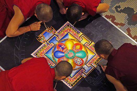 Overhead view of four people gathered around an ornate colored mandala sand painting on dark floor mats.