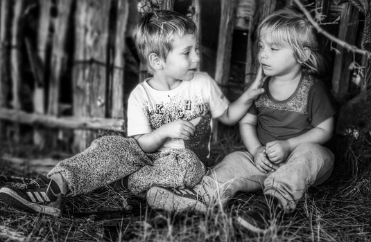 Two young children sitting together outdoors, facing each other in conversation among overgrown vegetation.