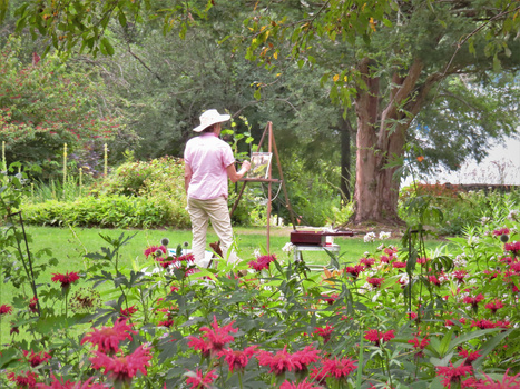 Gardener in pink shirt and white hat working near vibrant red flowers in a lush garden with mature trees.