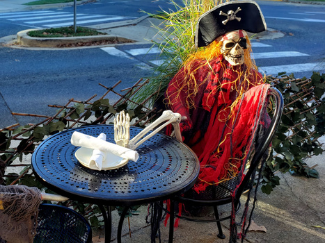 Skeleton figure in red coat and pirate hat seated at blue outdoor table with white plate and utensils.