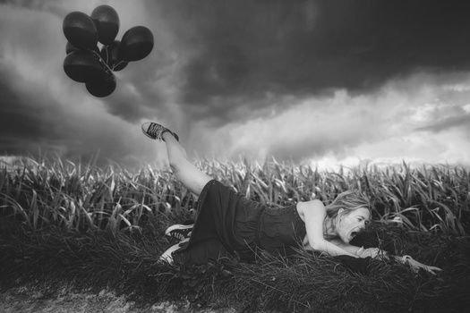 Person lying in a cornfield holding balloons against a dramatic cloudy sky.