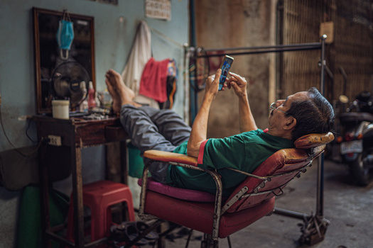 Man reclined in chair holding smartphone with tools and workspace visible behind him.