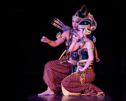 Two performers in ornate traditional costume with elaborate headdresses and beaded clothing, posed on a dark stage.
