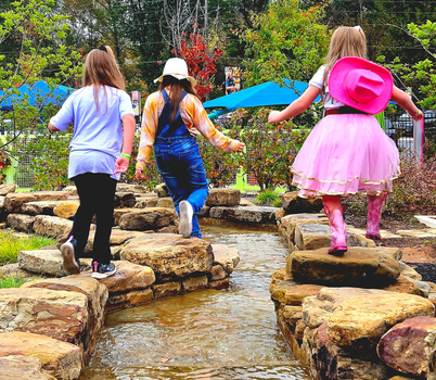 Three children crossing a shallow stream on stepping stones in a garden setting.