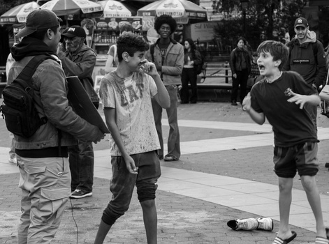 Black and white street photography of children playing in an outdoor public space with market umbrellas and crowds in the background.