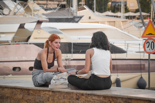 Two women sitting on a waterfront ledge with boats docked in the background.