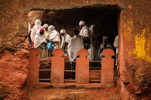 Pilgrims in white robes gather inside an ancient stone church with arched window openings.