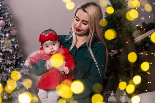 Woman in dark green cardigan holding infant in red dress and matching hat, surrounded by warm golden bokeh lights and decorated evergreen trees.