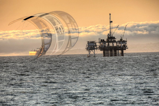 Offshore oil platform with billowing smoke beside a sailing vessel with large white sails at golden hour.