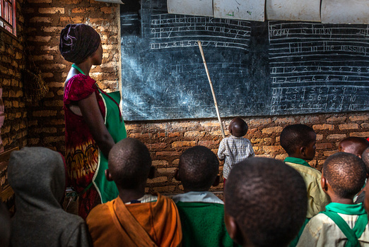 Teacher pointing at chalkboard while students sit facing away from camera in a brick classroom.