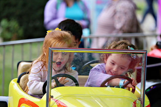 Young children with blonde hair laughing in a yellow and red amusement park ride with protective barriers.