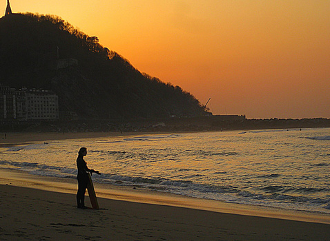 Silhouetted surfer carrying a surfboard walking into golden-hour ocean waves.
