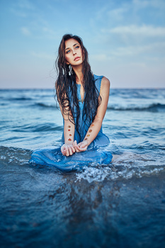 Woman kneeling in shallow ocean water wearing a wet blue dress, looking at camera.