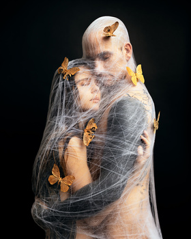 Person wrapped in spider web with yellow and orange butterflies in studio portrait against black background.