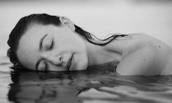 Woman's face and shoulder resting on water surface with eyes closed in monochrome.