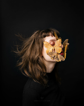 Woman with long brown hair holding a dried yellow leaf in front of her face against a black background.