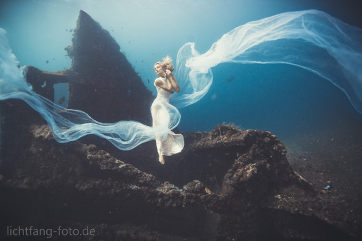 Woman in flowing white dress and veil suspended underwater among rocky formations with blue water above.
