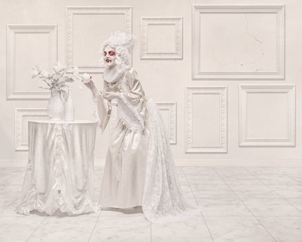 Elaborately costumed figure in white lace and skull makeup stands beside a round table with flowers in a minimalist interior.