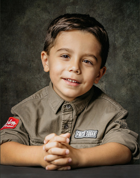 Young boy in scout uniform smiling at camera with hands clasped.