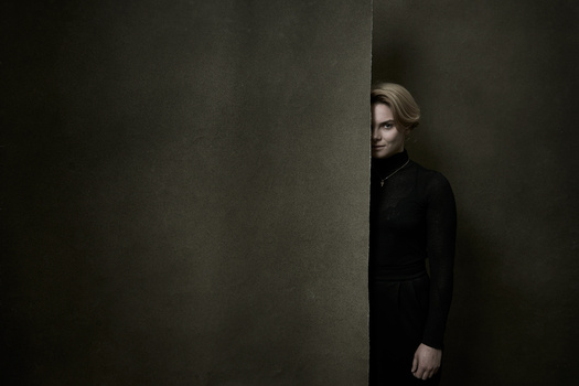 Person in black turtleneck standing partially hidden behind a large neutral-toned architectural column.