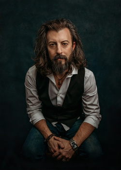 Man with long dark hair and beard wearing a black vest over a white shirt against a dark teal background.