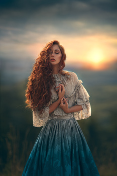 Woman with long curly hair wearing a lace blouse and teal skirt gazing upward against a dramatic sunset sky.