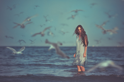 Woman in white dress standing in shallow ocean water with birds flying overhead.