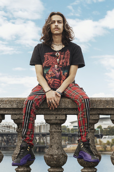 Person with long hair seated on stone balustrade wearing graphic band tee and tartan pants against cloudy sky.