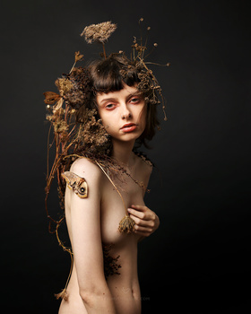 Young person with short dark hair and bangs, posed against a dark background with dried botanical elements arranged around their head and shoulders.
