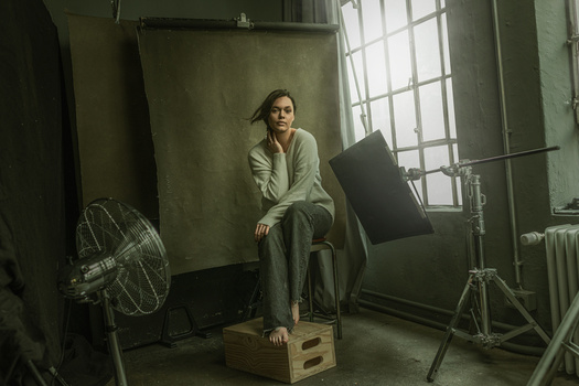 Woman sitting on wooden crate in studio with professional lighting setup and backdrop.