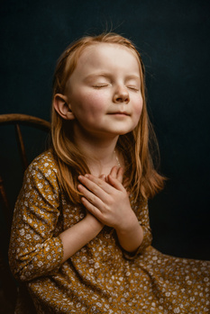 Young girl with reddish-brown hair wearing a mustard floral dress, eyes closed with hand at neck against dark background.