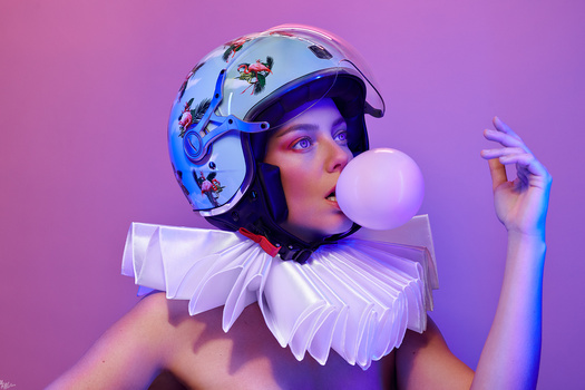 Person wearing a futuristic helmet and white ruffled collar, blowing a pink bubble against a purple background.