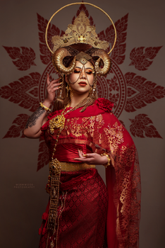 Woman in ornate red costume with gold crown and winged headdress against brown patterned backdrop.