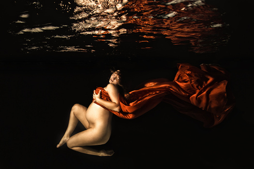 Nude figure suspended underwater with flowing orange fabric against dark water and light surface.