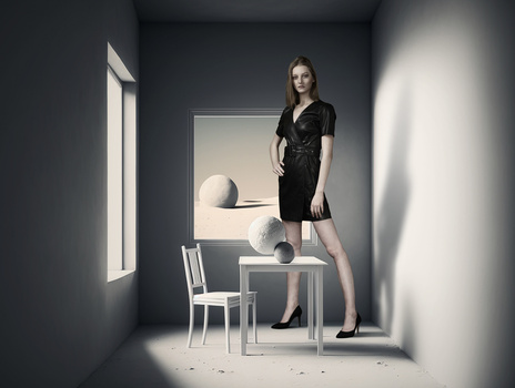 Woman in black wrap dress standing on white table in minimalist room with geometric still life artwork.