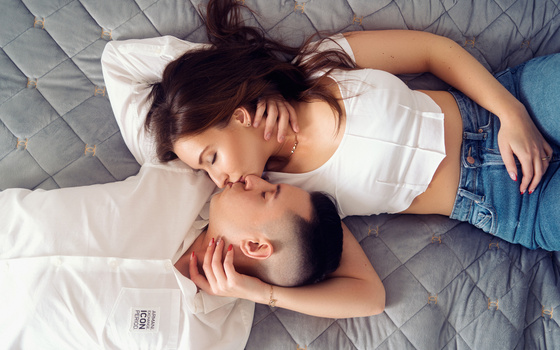 Overhead view of a couple lying on a quilted bed, heads close together and eyes closed in an intimate moment.