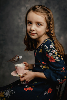 Young girl with braided hair wearing a dark floral dress, holding a dessert cup and looking at camera.