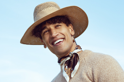 Smiling man wearing a tan cowboy hat and patterned bandana against a clear blue sky.