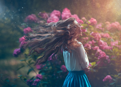 Woman with long flowing hair in blue dress standing among pink flowering bushes with golden sunlight.