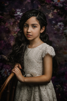 Young girl in a lace dress holding a wooden stick against a floral backdrop.