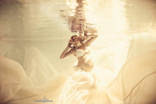 Woman submerged in water with arms raised, shot from below with bright sunlight filtering through.