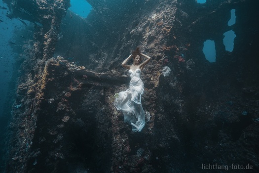 Freediver in white wetsuit suspended inside a dark shipwreck with deteriorated metal walls and openings to blue water above.