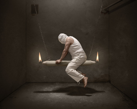 Man in white clothing suspended on a swing in a dark concrete room with dramatic side lighting.