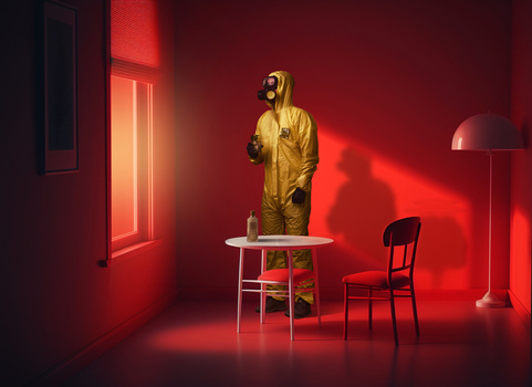 Figure in yellow hazmat suit standing on table in monochromatic red room with chair and lamp.