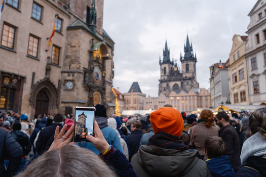 Crowded town square with person holding smartphone, Gothic church towers visible in background.