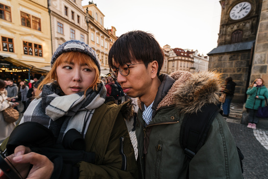 Two people wearing winter coats and scarves stand together in a European town square near a clock tower.