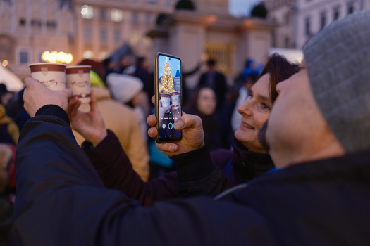 Person holding smartphone capturing video of illuminated architectural monument at dusk in crowded urban plaza.
