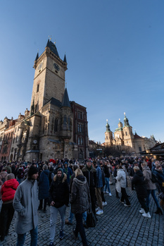 Tall Gothic tower with clock face rising above a crowded town square under clear blue sky.